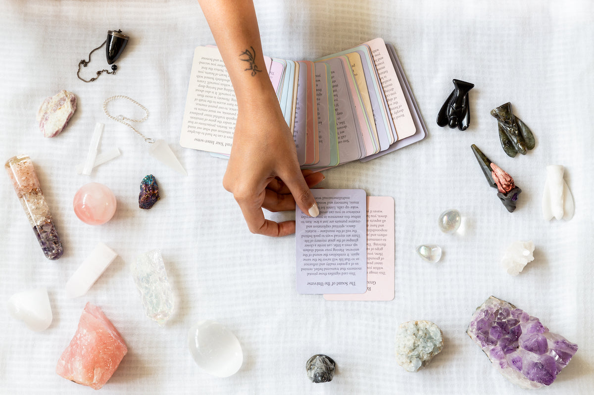 Gifts collection banner for The Tiny Topaz showing a hand laying down a tarot card on a table with surrounding crystals and angel stones.