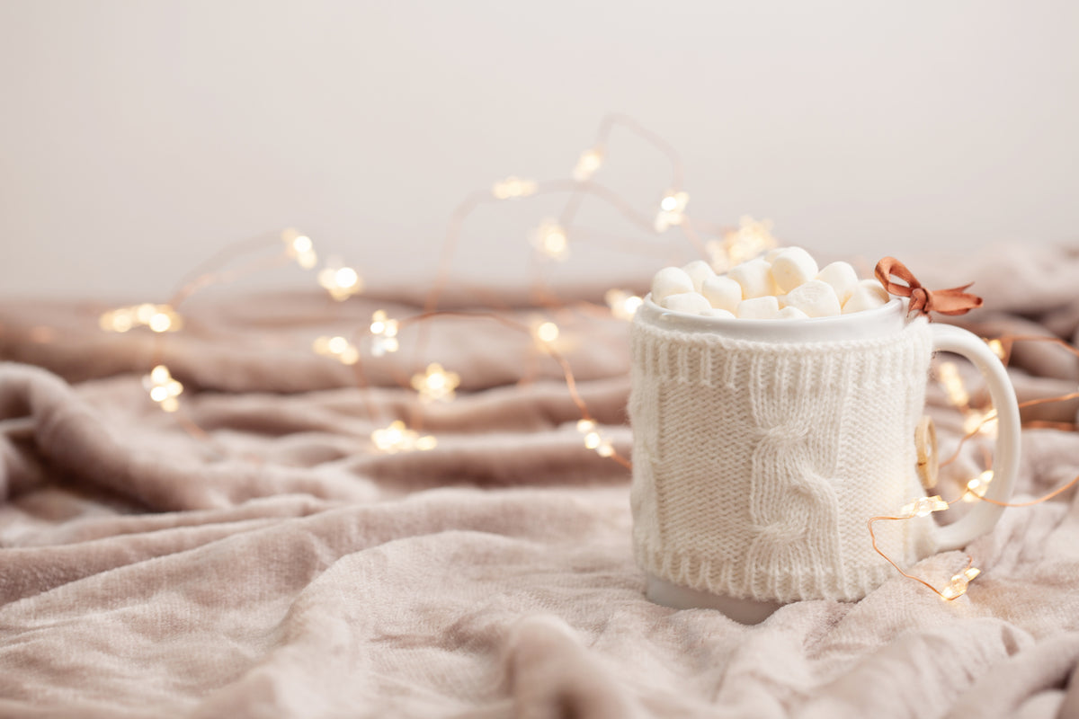 Mugs and glasses collection banner for The Tiny Topaz showing a cozy blanket and fairy lights, with a mug in a knitted cover that is full of marshmallows.